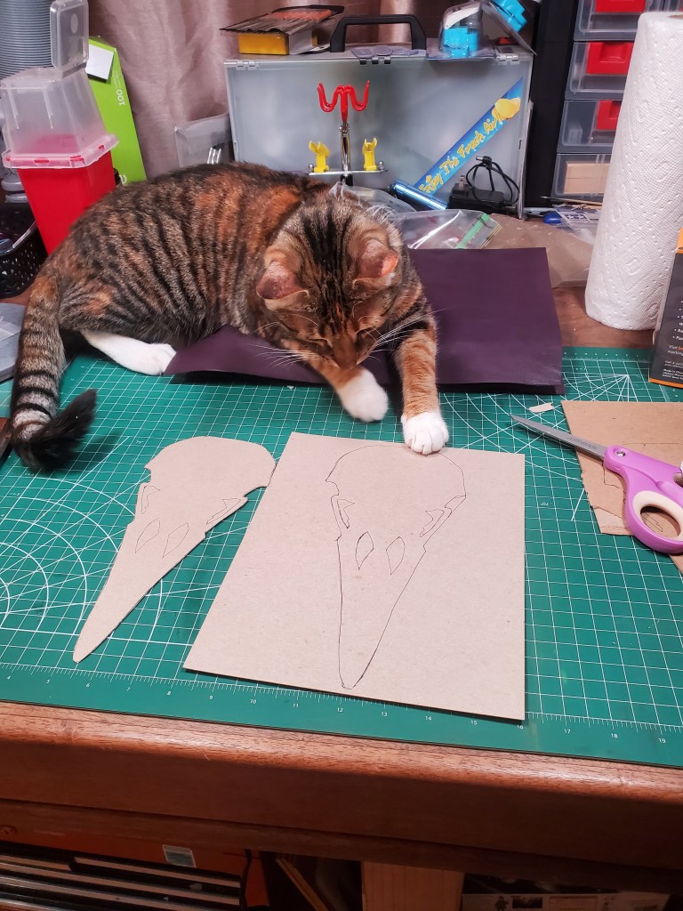 Drawing of a raven skull on thin card board. A second drawing of another layer of it cut out. A torbie cat named Sweet Potato examines the work.
